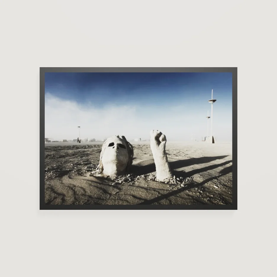Eye-level view of a framed photograph of a sculpted face and fist rising from the playa, hung on a white wall