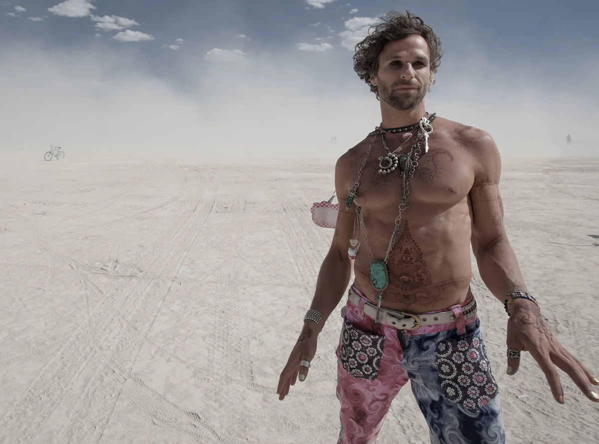 A shirtless man with necklaces and patterned pants standing in the dust